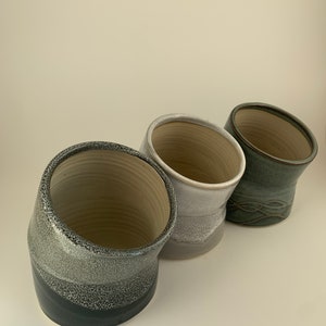 Peut inclure: Trois vases en céramique dans les tons de gris et de vert. Les vases ont un design angulaire unique. Le premier vase est gris foncé à la base, s'estompant vers un gris plus clair avec une texture mouchetée. Le second est gris clair. Le troisième est vert avec un motif texturé.