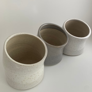 Peut inclure: Trois tasses en céramique avec un glaçage moucheté. Les tasses sont alignées, la tasse blanche à l'avant, la tasse grise au milieu et la tasse blanche à l'arrière.