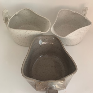 Puede incluir: Tres cuencos de cerámica con esmalte moteado. Dos cuencos son blancos con motas grises y un cuenco es gris con motas blancas. Los tres cuencos tienen un asa.