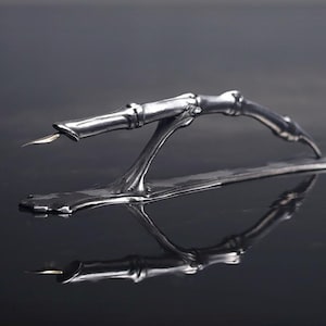 May include: A silver-toned letter opener with a unique skeletal hand design. The opener features a blade extending from the index finger, and rests on a rectangular base. The object is highly reflective, with a dark background.