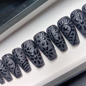 May include: A set of ten matte gray coffin-shaped press-on nails. Each nail features a raised, glossy black floral and geometric design. The nails are arranged in a row inside a white box.