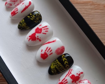 May include: A set of ten press-on nails featuring a crime scene theme. The nails alternate between a white base with red blood splatter and handprint designs, and a black base with yellow police caution tape patterns and the text "POLICE LINE".