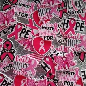 May include: A collection of pink and white stickers with various designs and text. The stickers feature pink ribbons, hearts, and butterflies, with phrases like "Believe," "Hope," and "A Cure Worth Fighting For." The background is a sparkly silver.