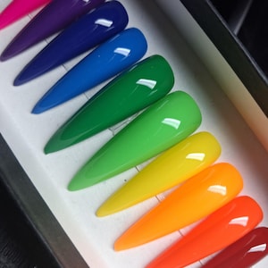 May include: A set of ten stiletto-shaped press-on nails arranged in a rainbow gradient. The colors include hot pink, purple, dark blue, bright blue, dark green, light green, yellow, orange, bright orange, and deep red with a glossy finish.
