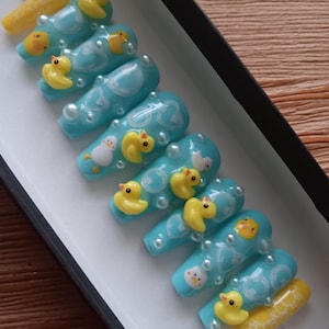 May include: A set of press-on nails with a light blue base, decorated with white cloud designs, yellow rubber duckies, and small pearl accents. One nail is yellow with a white pattern. The nails are displayed in a black box.