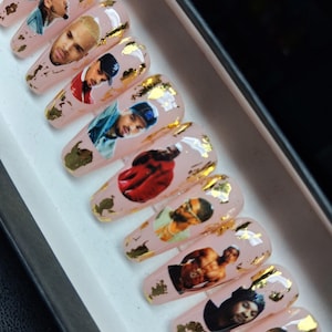 May include: Set of artificial nails with a light pink base, featuring images of a celebrity and gold foil accents. The nails are long and coffin-shaped, with a glossy finish. The images are in various poses and outfits.