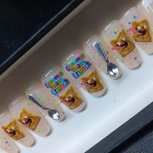 May include: A set of press-on nails with a clear base, decorated with colorful confetti and cartoon images of Cinnamon Toast Crunch cereal. Each nail features a smiling cereal character and a tiny silver spoon.