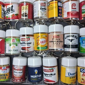 May include: A collection of miniature spice and seasoning jars, each with a white lid and a silver keychain. The jars feature labels with various brand names and seasoning types, including pepper, salt, and garlic. The arrangement is displayed in a clear plastic container.