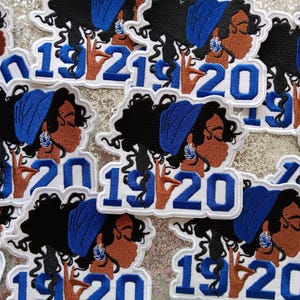 May include: Multiple white patches with blue and black embroidery. Each patch features a woman with blue hair and a blue headband, and the text "1920".
