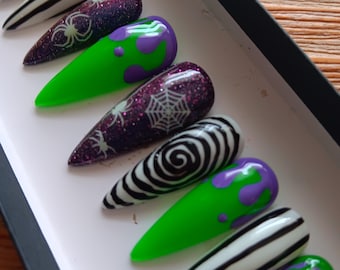 May include: A set of ten stiletto-shaped press-on nails featuring various designs including black and white spiral patterns, black and white stripes, purple glitter with white spider and web motifs, and bright green with purple slime drip accents.