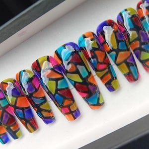 May include: Coffin-shaped press-on nails with a stained glass design. Each nail features a mosaic of colorful geometric shapes in shades of red, orange, yellow, green, blue, and purple, outlined in black. A white, abstract design is on each nail.