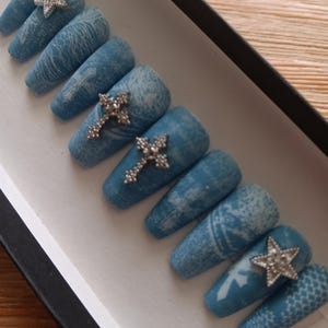 May include: A set of blue press-on nails with a textured design and silver embellishments. The nails feature a mix of patterns, including a cross and star, and are displayed in a black and white box. The nails are long and coffin-shaped.