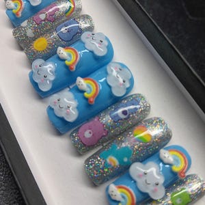May include: A set of ten press-on nails with a blue base and glitter. Each nail features a different Care Bear character, a rainbow, and a cloud with a smiley face.