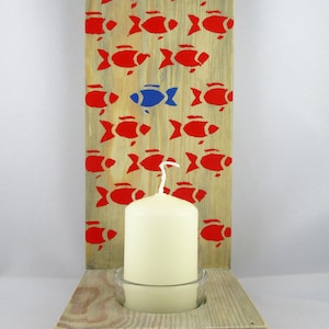 Può includere: Un portacandele in legno con una singola candela in un portacandele di vetro. Il portacandele è decorato con un motivo di pesci rossi, con un pesce blu al centro.