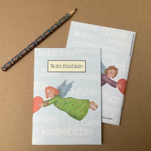Könnte beinhalten: Zwei Notizbücher mit Engel-Illustrationen und einem dekorativen Bleistift. Die Notizbücher haben einen hellblauen Einband mit dem deutschen Text "Notiz-Büchlein" und anderen Wörtern. Der Bleistift ist dunkelblau mit orangefarbenen und weißen Blumenmustern.