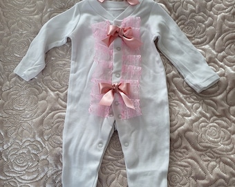 May include: White baby onesie with pink lace and satin bows. The onesie has long sleeves and snaps at the crotch. A matching pink satin headband with a bow is included.