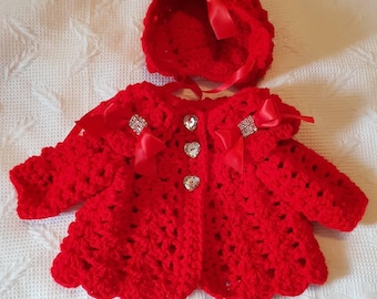 Puede incluir: Un cárdigan de bebé de punto rojo con un gorro a juego. Ambas piezas tienen botones en forma de corazón y adornos de pedrería en los lazos.