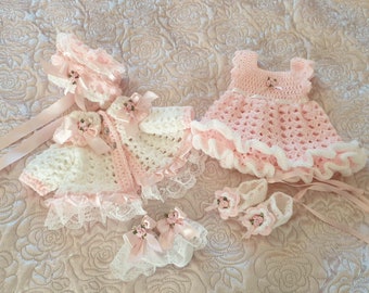 May include: A set of crocheted baby clothes in pink and white, including a dress, a cardigan, a hat, and booties. The set is decorated with lace and pink satin ribbons.