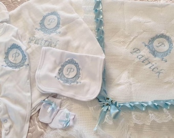 May include: A white baby outfit set with blue embroidery. The set includes a onesie, a burp cloth, a pair of booties, and a blanket. The blanket has a blue satin ribbon trim and a feather trim. The set is personalized with the name "Patrick" and the letter "P" in a decorative frame.