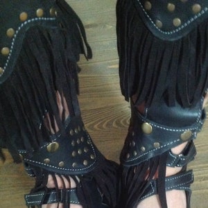 Puede incluir: Sandalias de cuero negro con flecos y tachuelas doradas. Las sandalias tienen un estilo gladiador con correas que suben por la pierna.