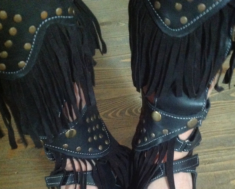 Puede incluir: Sandalias de cuero negro con flecos y tachuelas doradas. Las sandalias tienen un estilo gladiador con correas que suben por la pierna.