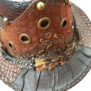 May include: A brown leather cowboy hat with a wide brim, decorated with feathers, metal accents, and a snakeskin pattern.