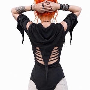 May include: Black, distressed-style top with cut-out back detail. The top features a unique design with cut-out sleeves and a long, pointed tail in the back. The model has orange hair and is wearing bracelets and rings.