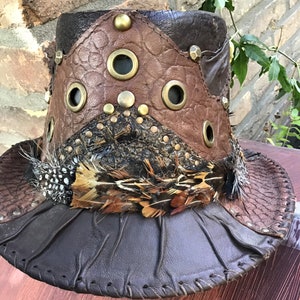 May include: A brown leather hat with a wide brim, decorated with feathers and metal accents. The hat has a unique design with multiple panels of leather sewn together.