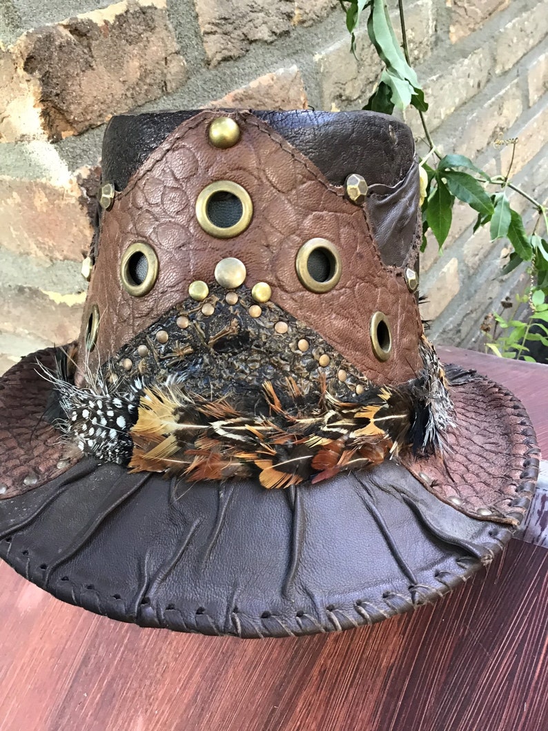 May include: A brown leather hat with a wide brim, decorated with feathers and metal accents. The hat has a unique design with multiple panels of leather sewn together.