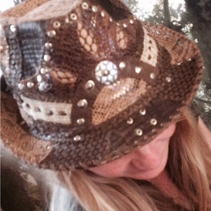 May include: A brown and beige cowboy hat with a lace design and silver embellishments. The hat has a wide brim and is decorated with rhinestones.