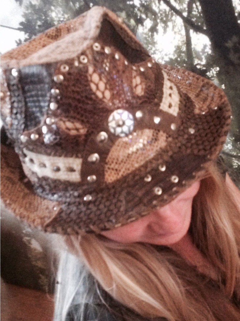 May include: A brown and beige cowboy hat with a lace design and silver embellishments. The hat has a wide brim and is decorated with rhinestones.