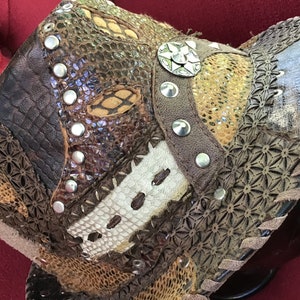 May include: A brown leather hat featuring a mix of textures and patterns, including snakeskin, floral, and geometric designs. The hat is decorated with silver studs and a large silver star-shaped embellishment.