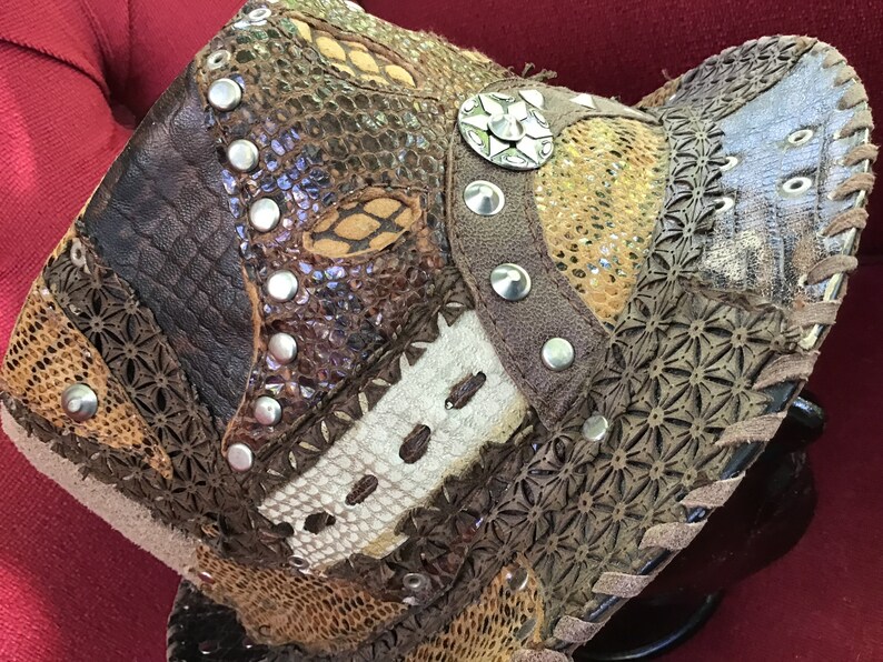 May include: A brown leather hat featuring a mix of textures and patterns, including snakeskin, floral, and geometric designs. The hat is decorated with silver studs and a large silver star-shaped embellishment.
