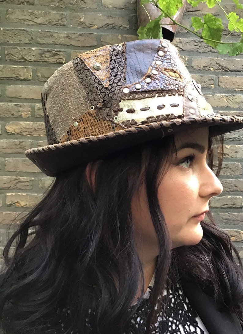 May include: A brown leather hat with a patchwork design. The hat is made of different types of leather, including suede, snakeskin, and embossed leather. The hat has a wide brim and a tall crown.