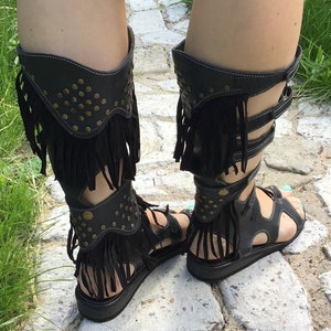Puede incluir: Sandalias gladiadoras de cuero negro con flecos y tachuelas doradas. Las sandalias tienen una suela gruesa y una correa que sube por la pierna.