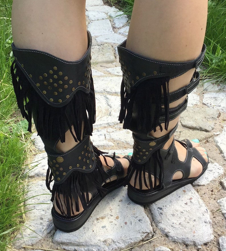 Puede incluir: Sandalias gladiadoras de cuero negro con flecos y tachuelas doradas. Las sandalias tienen una suela gruesa y una correa que sube por la pierna.