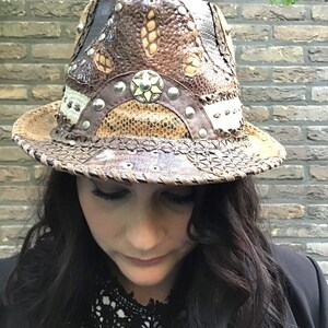 May include: A brown leather fedora hat with a snakeskin pattern and silver studs. The hat has a wide brim and a tall crown.