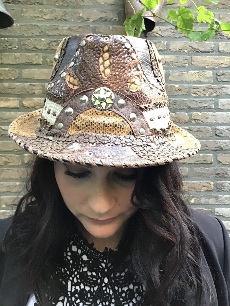 May include: A brown leather fedora hat with a snakeskin pattern and silver studs. The hat has a wide brim and a tall crown.
