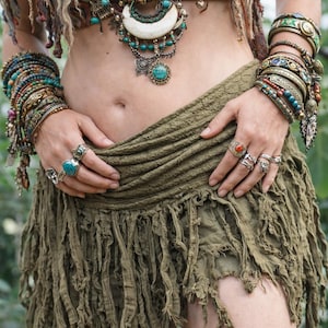 Puede incluir: Un conjunto de estilo bohemio con collares superpuestos, colgantes turquesas y múltiples pulseras y anillos. El atuendo incluye una falda con flecos y una blusa estampada, mostrando una estética de espíritu libre.