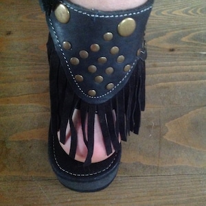 Puede incluir: Sandalias de cuero negro con flecos y tachuelas doradas. Las sandalias tienen una suela de plataforma gruesa.