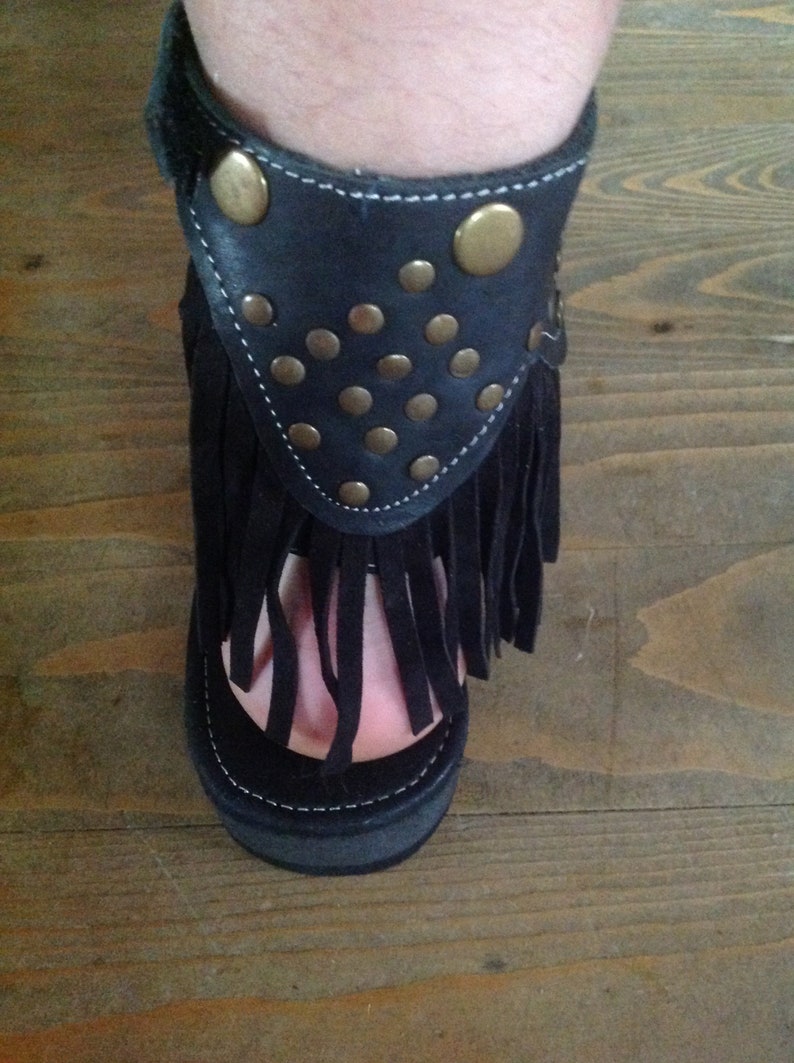 Puede incluir: Sandalias de cuero negro con flecos y tachuelas doradas. Las sandalias tienen una suela de plataforma gruesa.