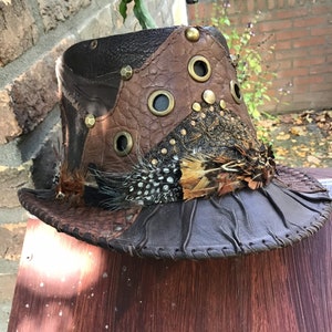 May include: A brown leather hat with feathers and metal accents. The hat has a wide brim and a tall crown. The hat is decorated with feathers, metal studs, and eyelets.