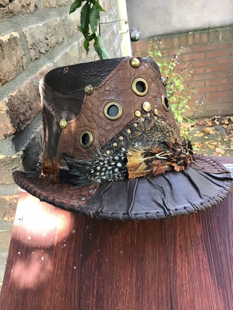 May include: A brown leather hat with feathers and metal accents. The hat has a wide brim and a tall crown. The hat is decorated with feathers, metal studs, and eyelets.