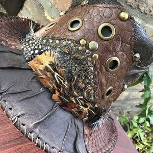 May include: A brown leather hat with a unique design featuring multiple textures, feathers, and metal accents. The hat has a wide brim and a pointed crown.