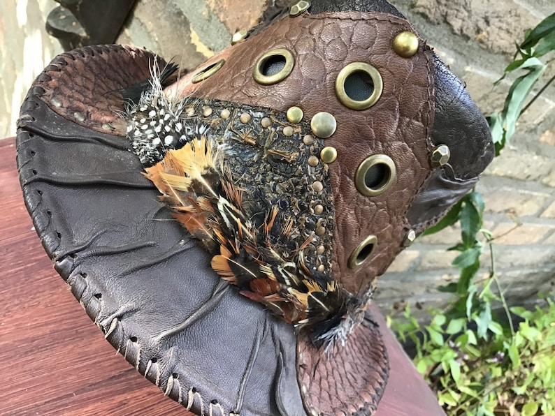 May include: A brown leather hat with a unique design featuring multiple textures, feathers, and metal accents. The hat has a wide brim and a pointed crown.