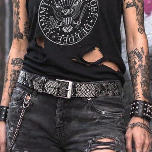 Op de afbeelding: Een zwart mouwloos shirt met versleten gaten en een witte cirkelvormige afbeelding met een adelaar en de tekst RAMONES JOHNNY JOEY DEE DEE, gecombineerd met een grijze en zwarte riem met slangenprint en een donkergrijze versleten spijkerbroek.