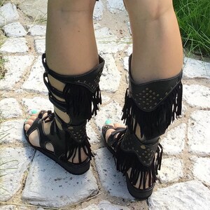 Puede incluir: Sandalias gladiadoras de cuero negro con flecos y detalles dorados. Las sandalias tienen una correa que sube por la pierna y se abrocha en la pantorrilla.