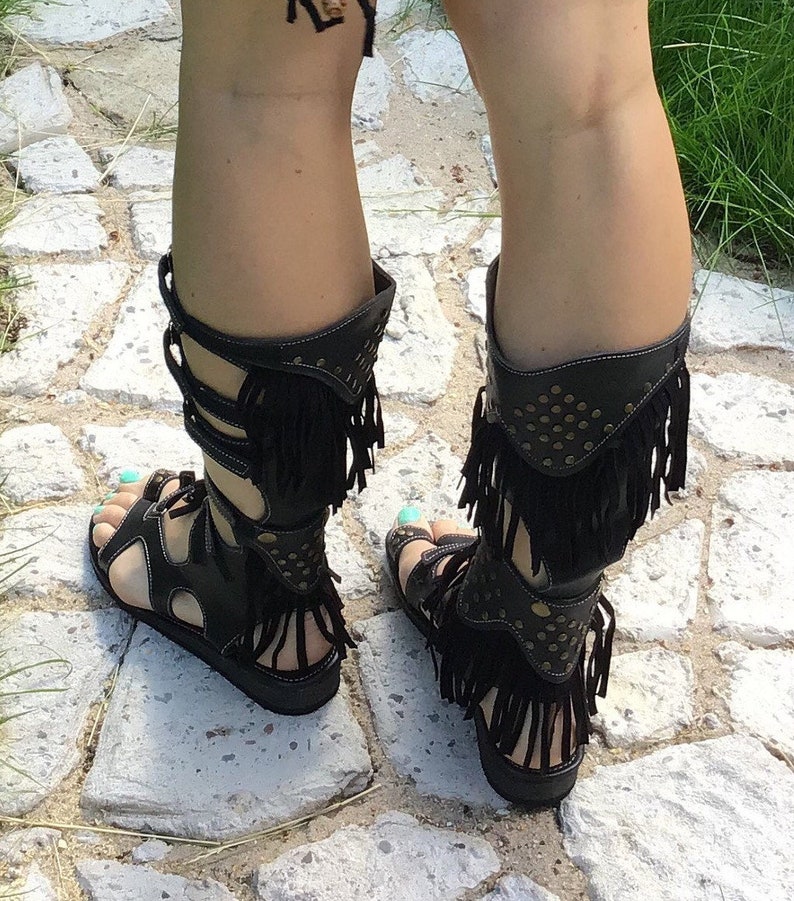 Puede incluir: Sandalias gladiadoras de cuero negro con flecos y detalles dorados. Las sandalias tienen una correa que sube por la pierna y se abrocha en la pantorrilla.