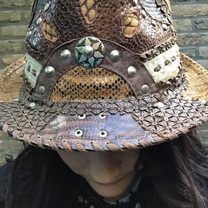 May include: A brown leather cowboy hat with a unique design featuring a variety of textures and embellishments, including metal studs and a large star-shaped button.