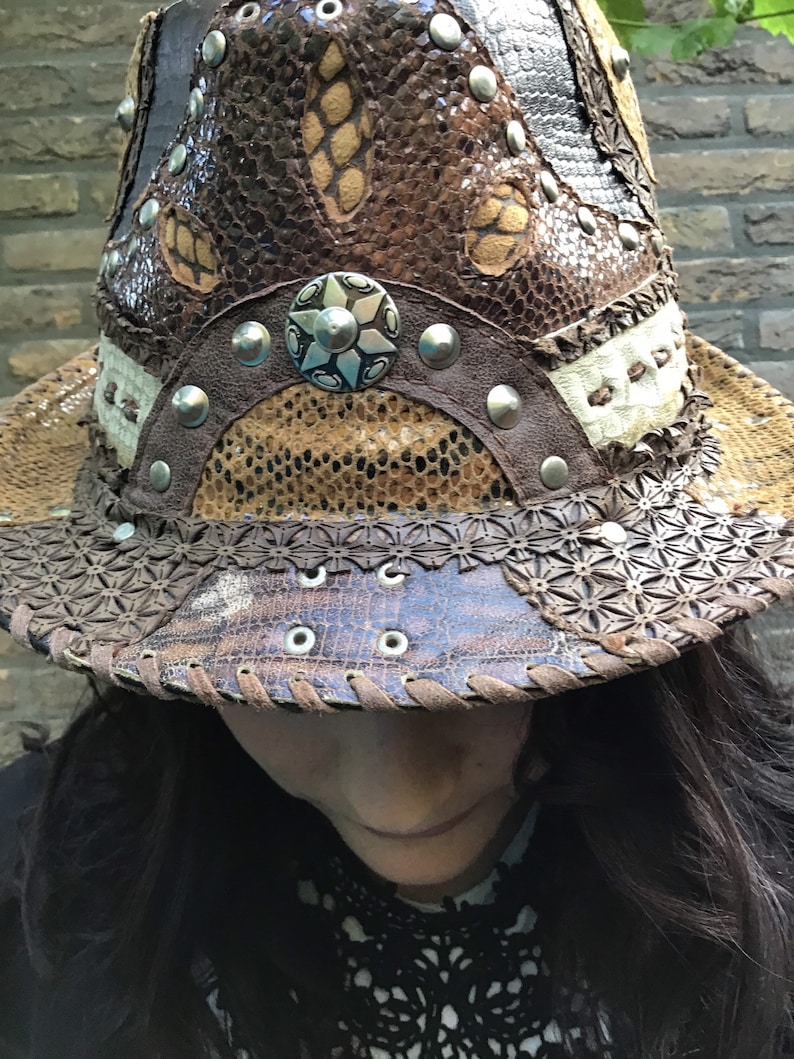 May include: A brown leather cowboy hat with a unique design featuring a variety of textures and embellishments, including metal studs and a large star-shaped button.
